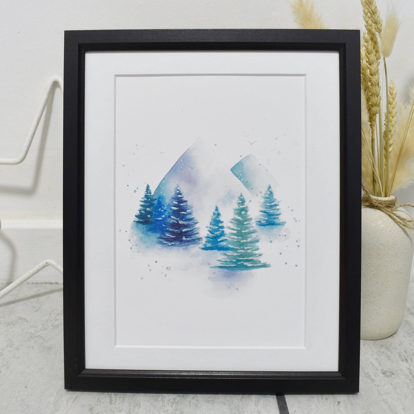 Framed artwork of blue trees on a white background, placed on a light wooden surface.