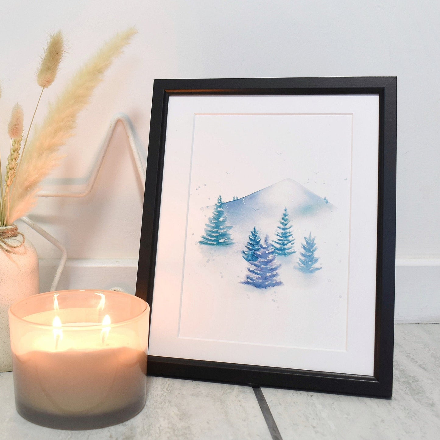 Framed artwork of trees and mountains next to a lit candle and vase with dried plants on a light wooden floor.
