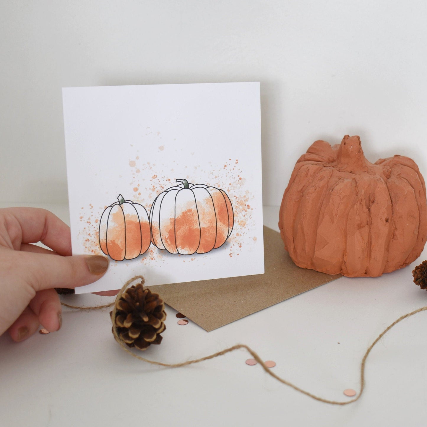 Hand holding a card with pumpkins and a small pumpkin decoration on a white surface.