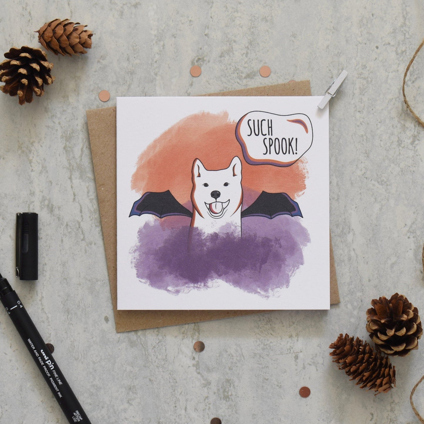 Halloween-themed greeting card with a dog wearing a bat costume on a textured surface with pine cones and a pen.