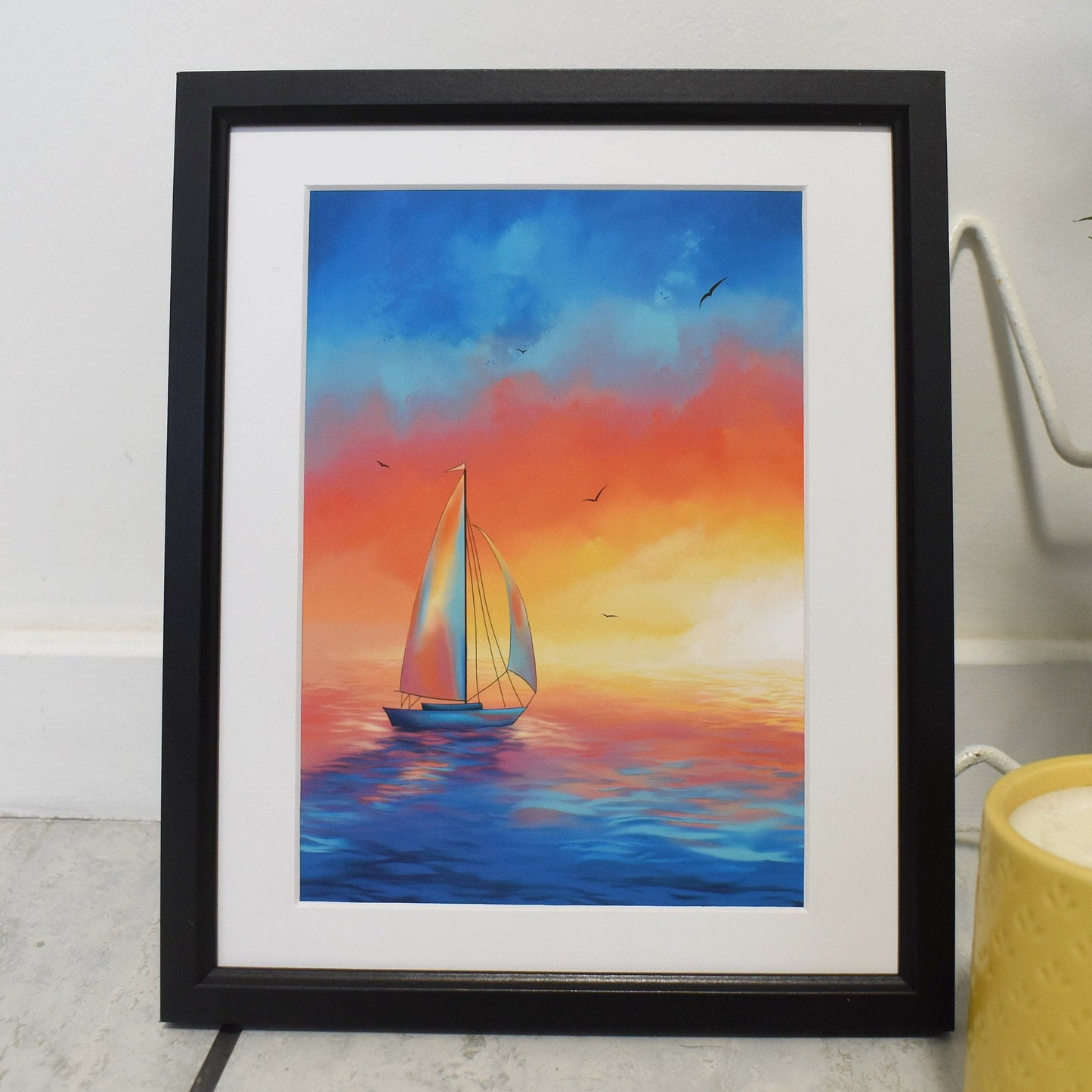 Framed painting of a sailboat at sunset with a candle and plant in the background.