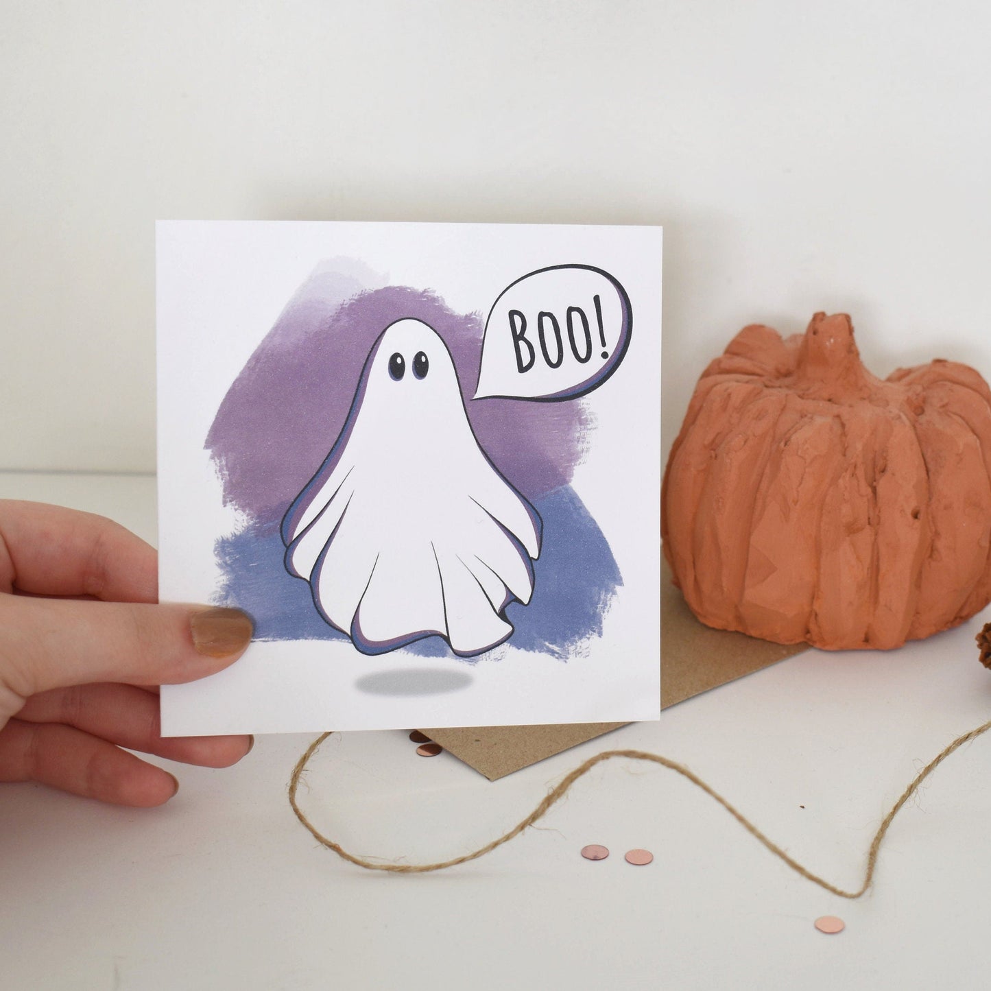 Hand holding a Halloween-themed card with a ghost and 'BOO!' text, placed on a surface with a pumpkin and pinecones.