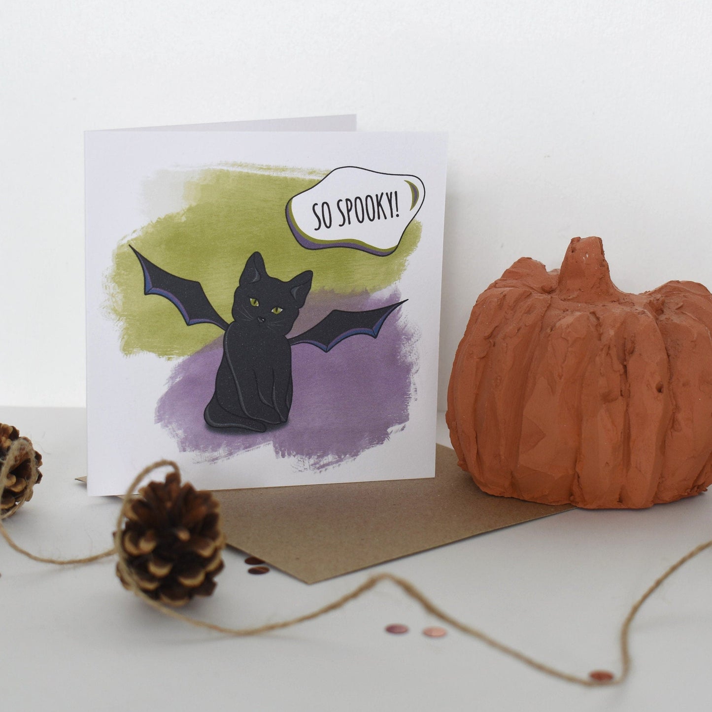 Halloween-themed card with a black cat and bat illustration, accompanied by a small pumpkin and pinecones on a white background.