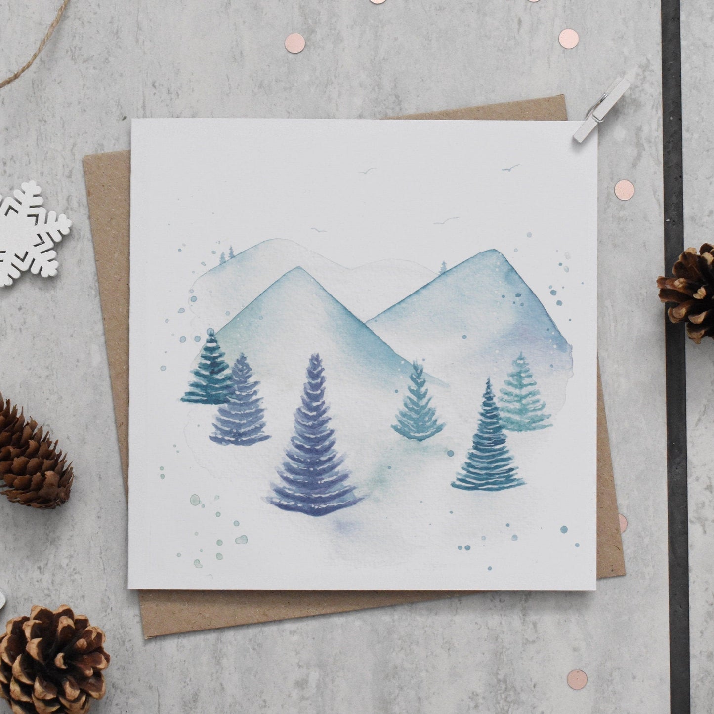 Greeting card with winter scene of trees and mountains on a gray surface with decorative elements.