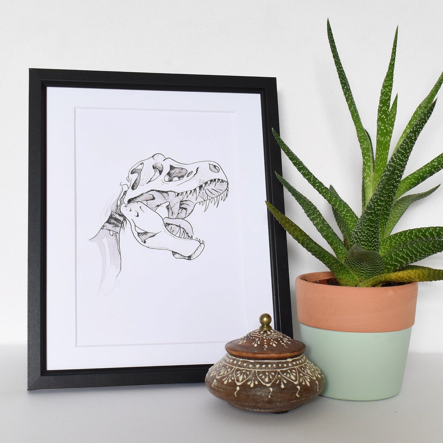 Framed artwork next to a potted plant on a white surface