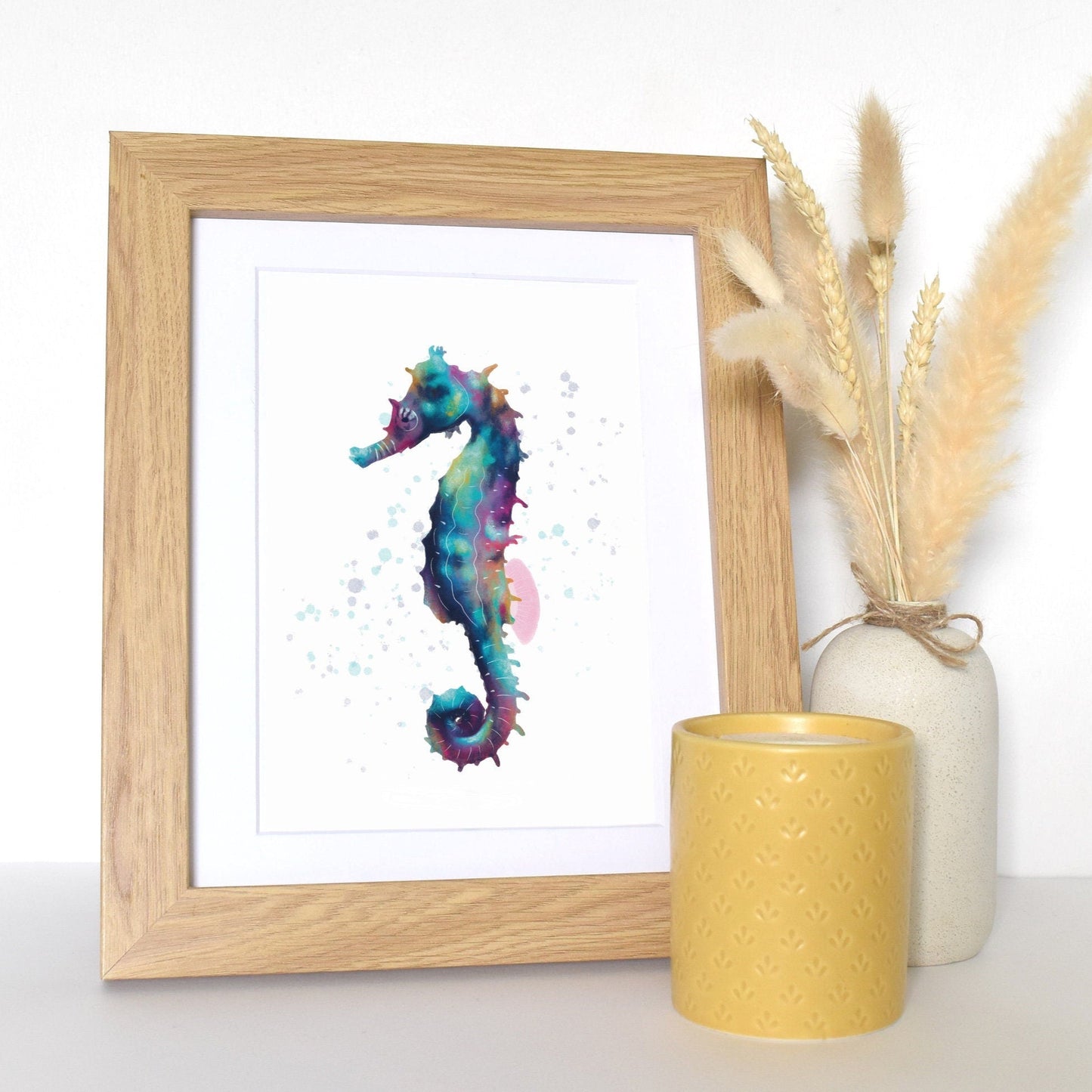 Framed artwork of a colorful seahorse next to a yellow candle and vase with dried plants on a white surface.