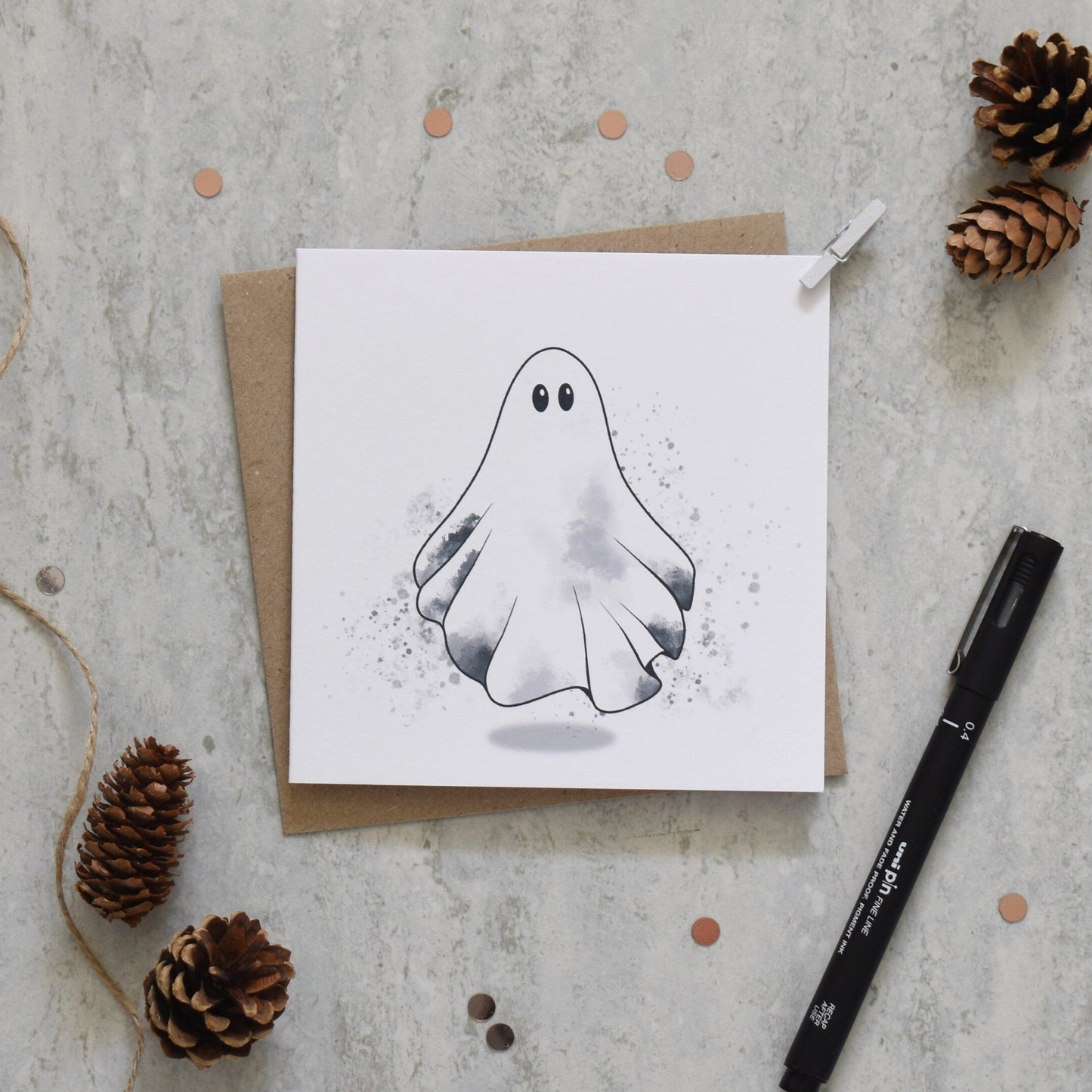 Greeting card with ghost illustration on a textured surface with pine cones and a pen.