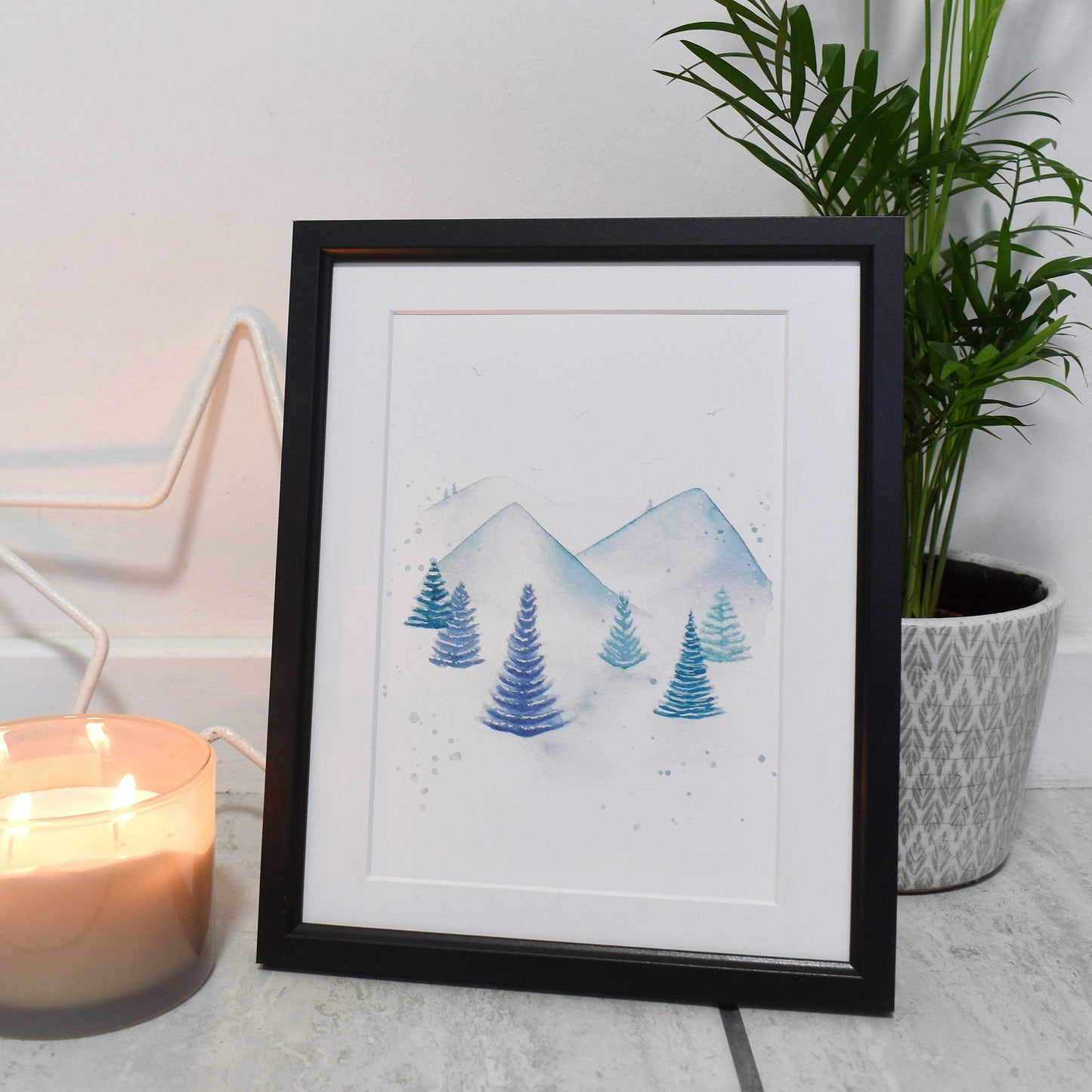 Framed artwork of mountains and trees next to a candle and plant on a light surface.