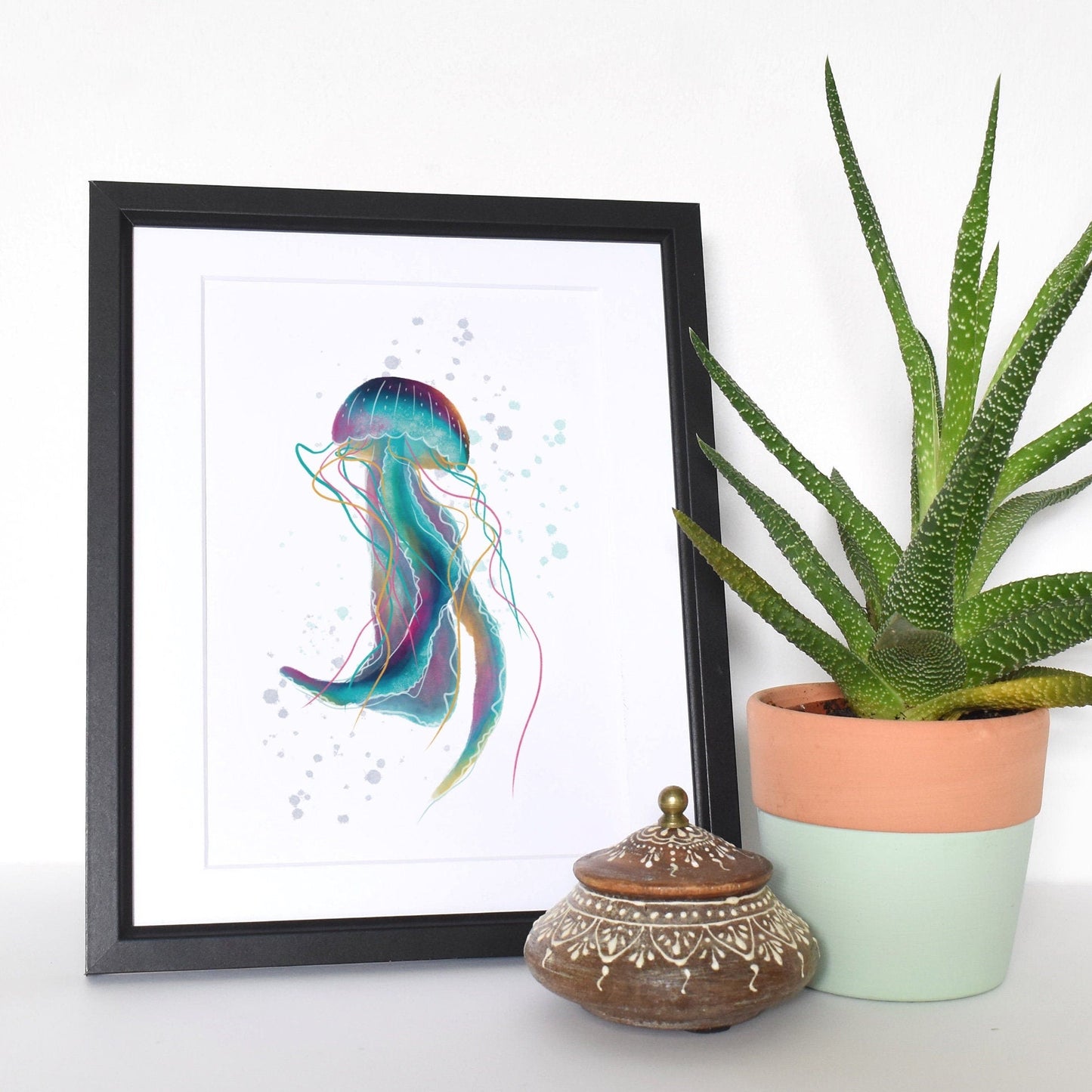 Framed artwork of a colorful jellyfish next to a potted plant on a white surface