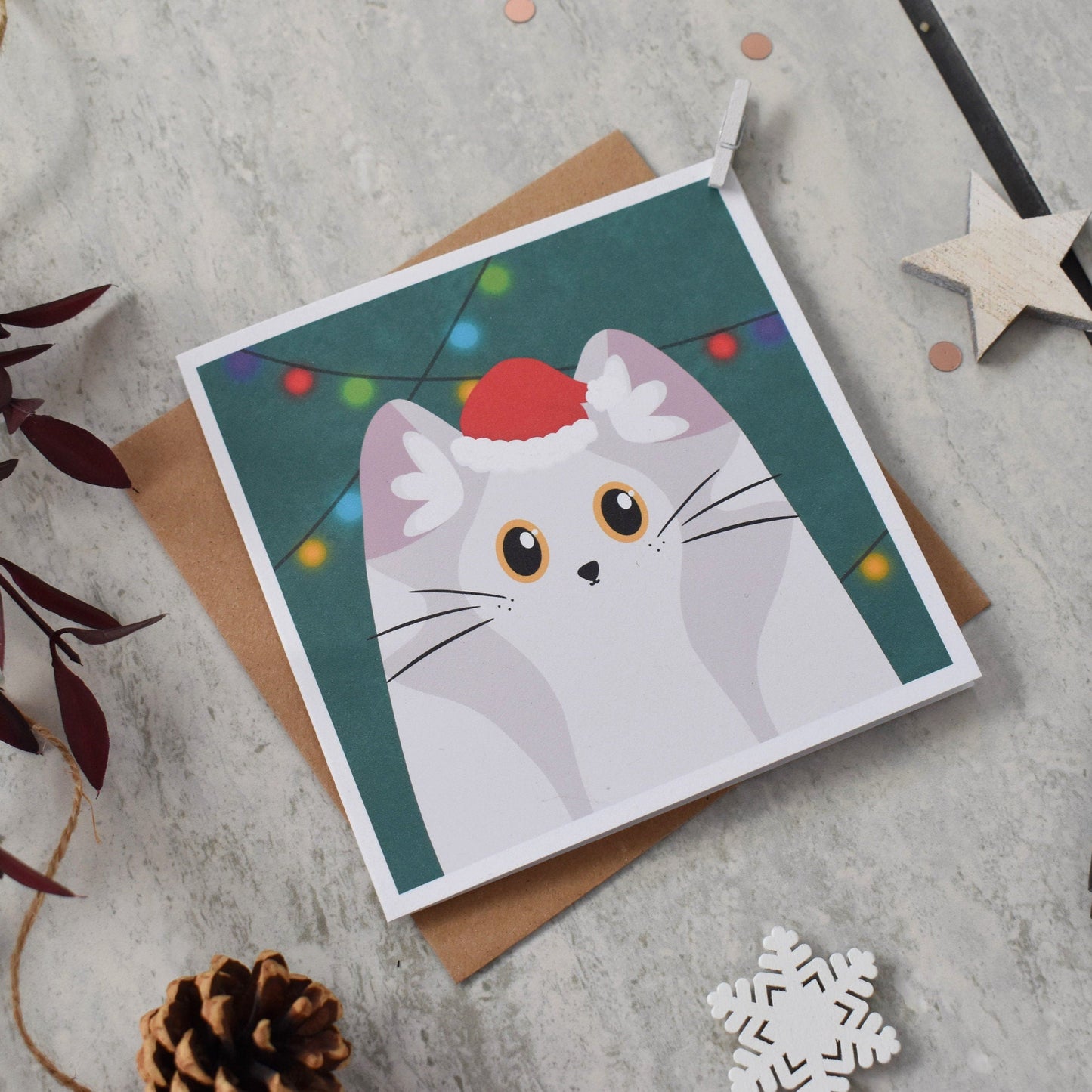 Card with a cat wearing a Santa hat and Christmas lights on a gray surface with decorative items.