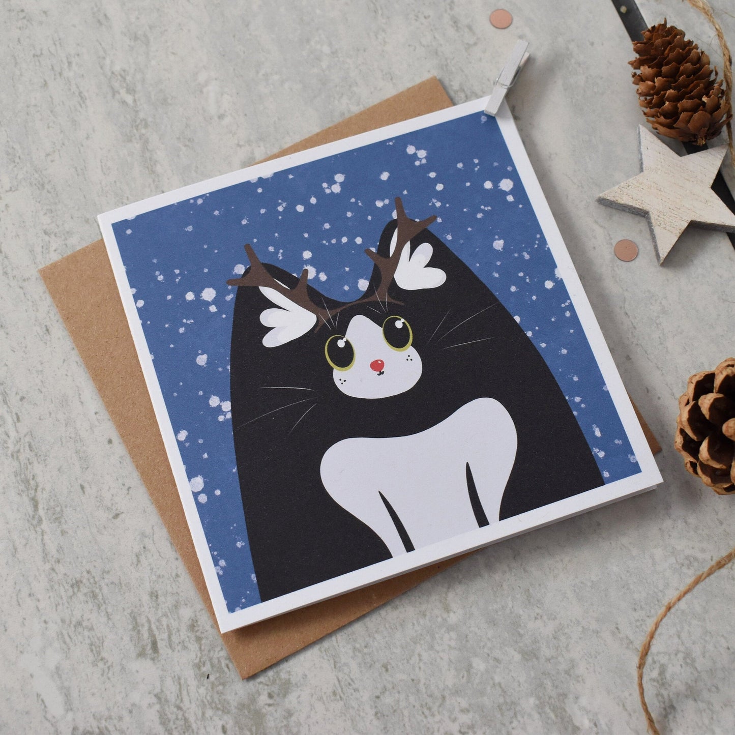 Greeting card with a black and white cat illustration wearing reindeer antlers on a blue snowy background, placed on a textured surface with decorative elements.