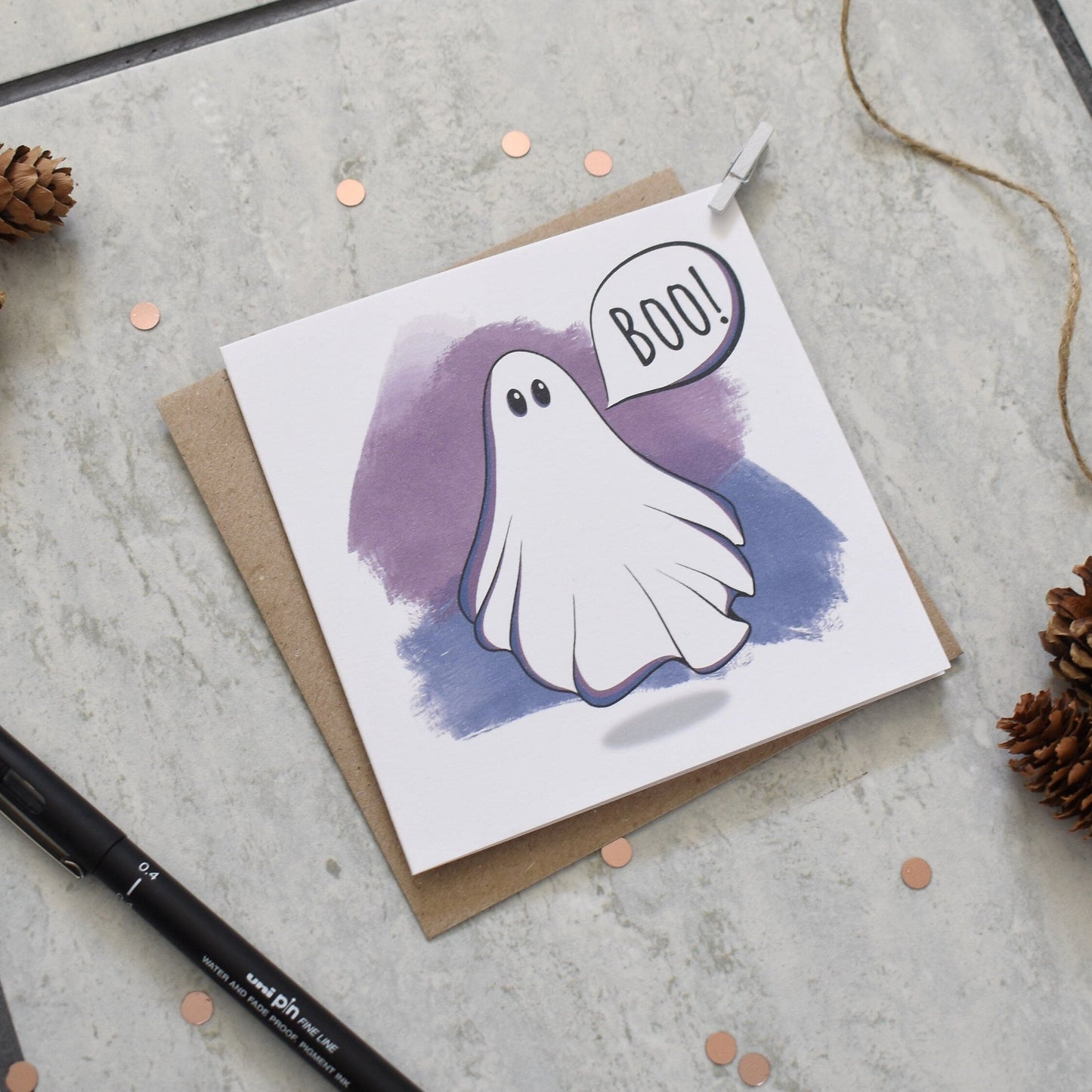 Greeting card with a ghost illustration and 'BOO!' text, surrounded by pine cones and a pen on a textured surface.