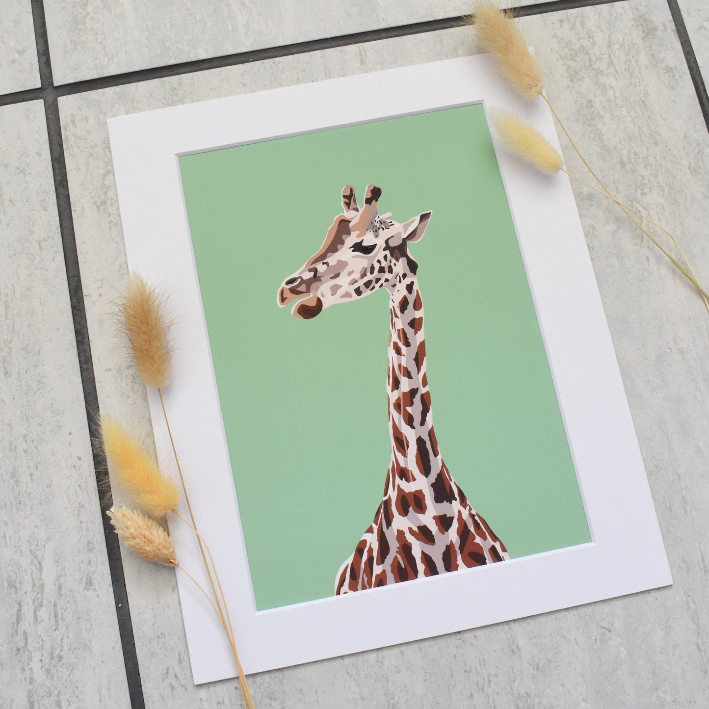 Framed artwork of a giraffe on a light green background with decorative elements on a tiled floor.