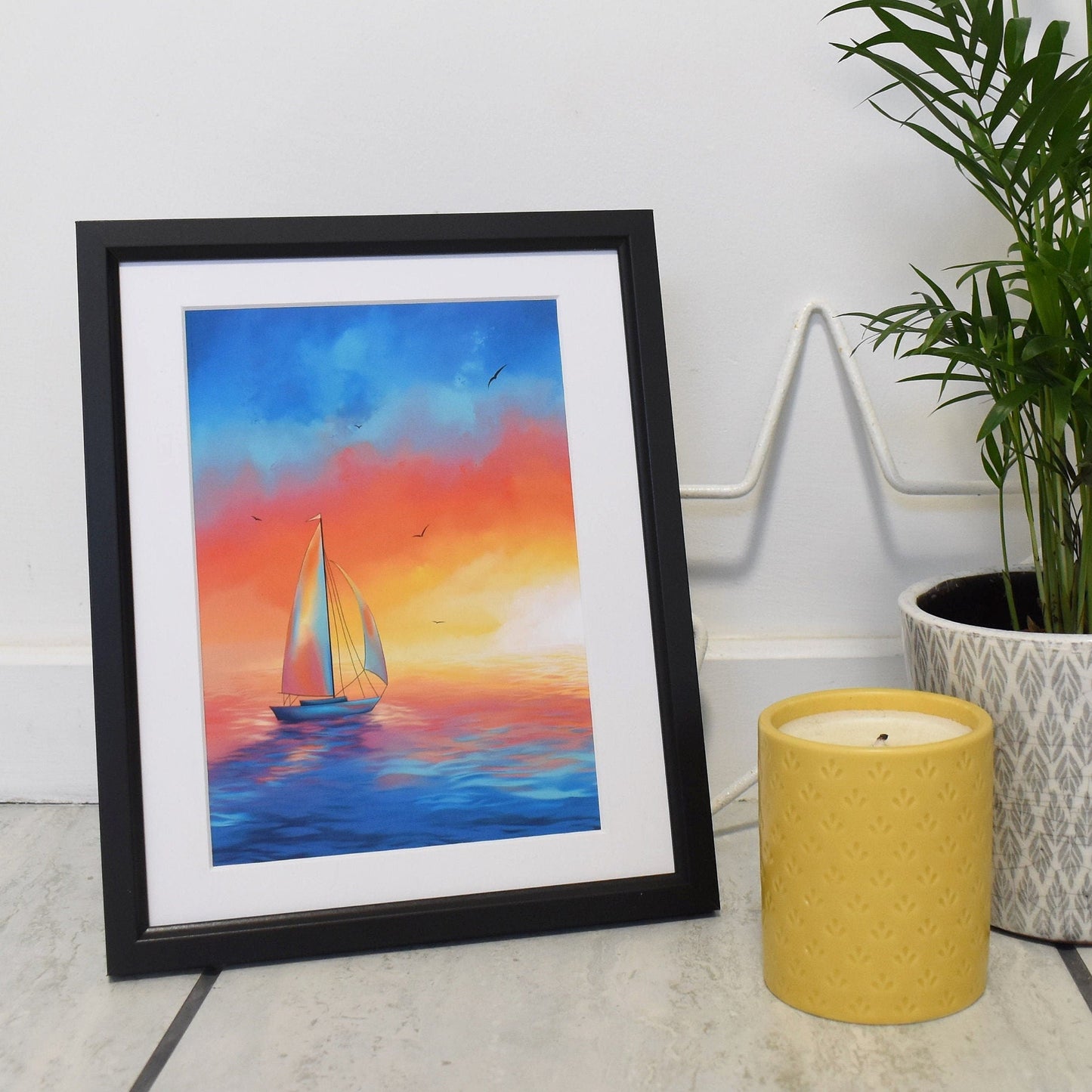 Framed artwork of a sailboat at sunset on a table with a candle and plant.