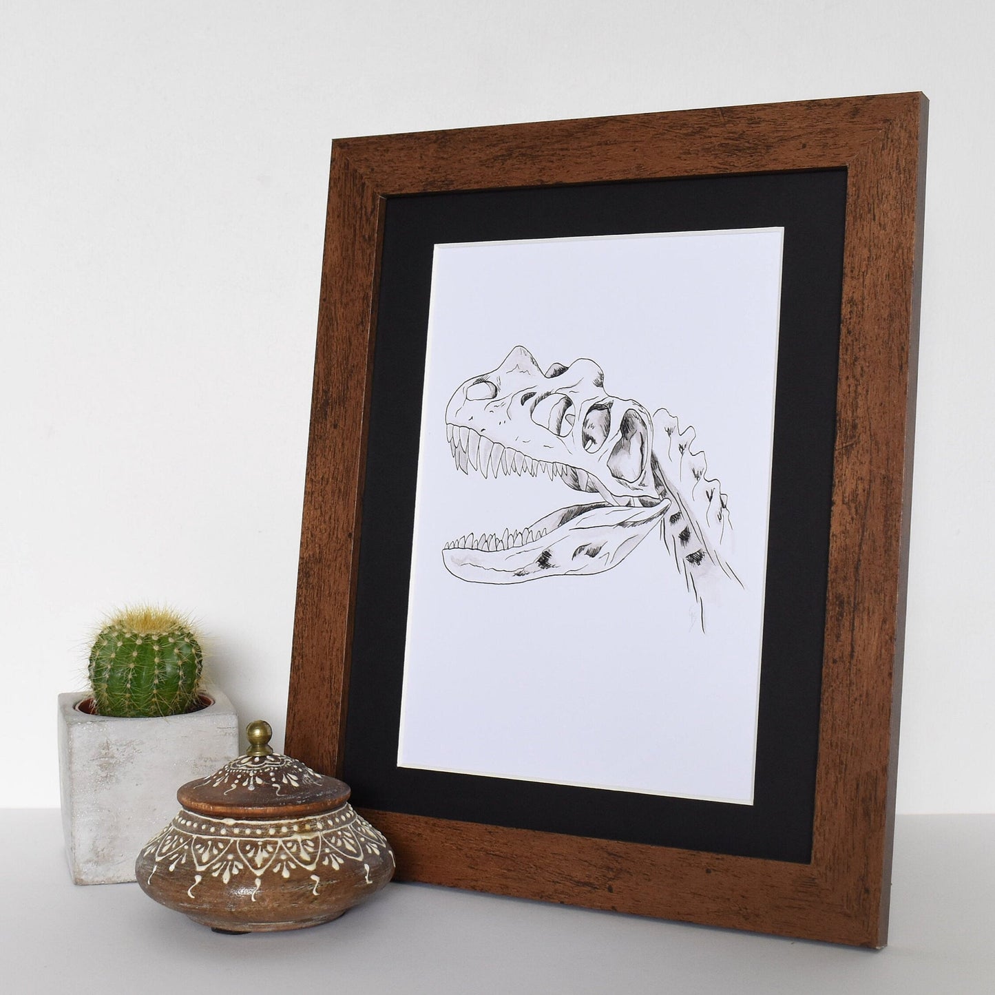Framed black and white illustration of a dinosaur skeleton on a white surface with decorative items.