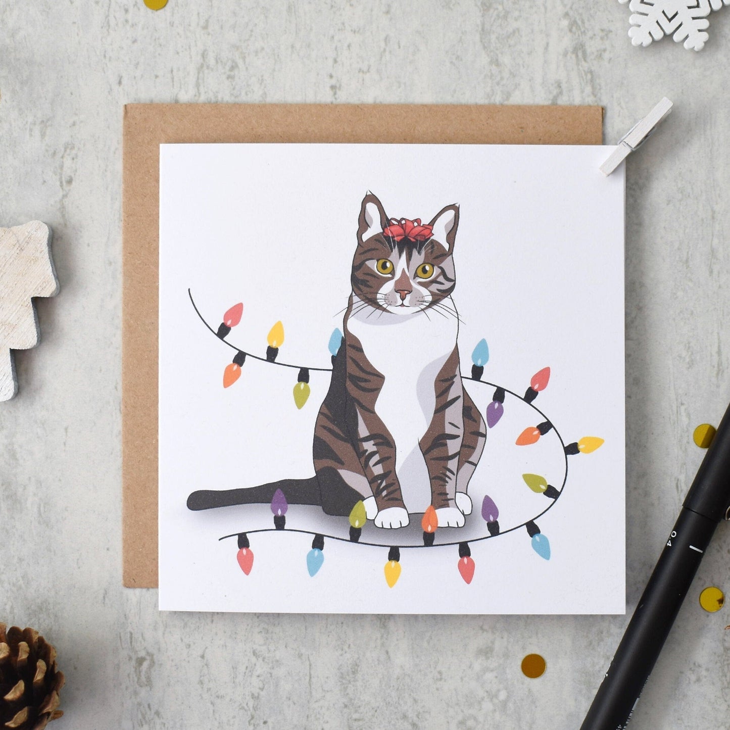 Card with a cat illustration on a string of lights, surrounded by decorative items on a gray surface.