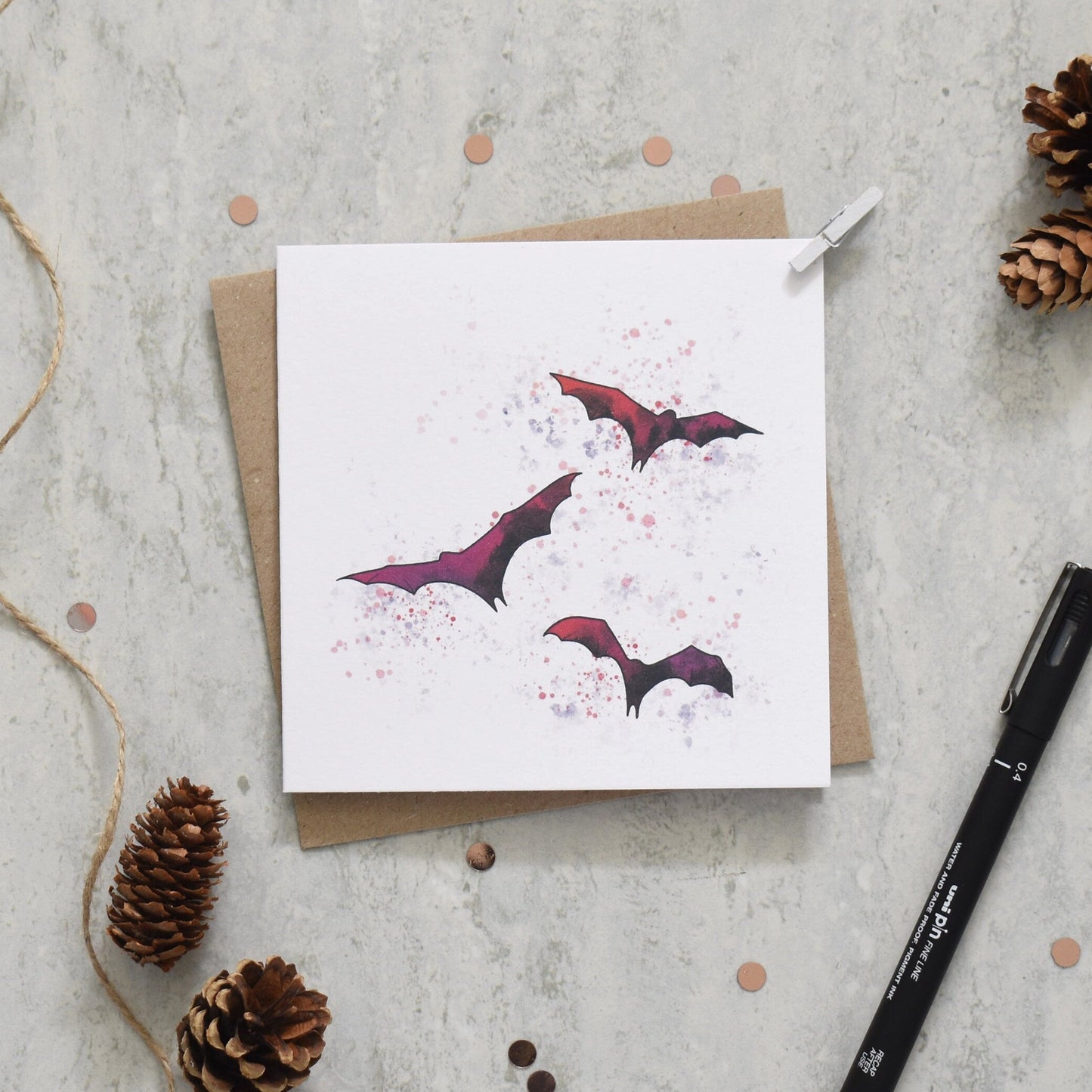 Greeting card with bat design on a textured surface with pine cones and a pen.