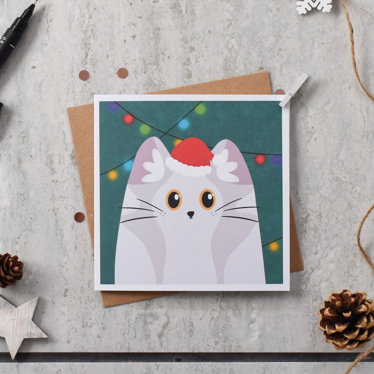 Card with a cat illustration wearing a Santa hat on a textured surface with decorative items.