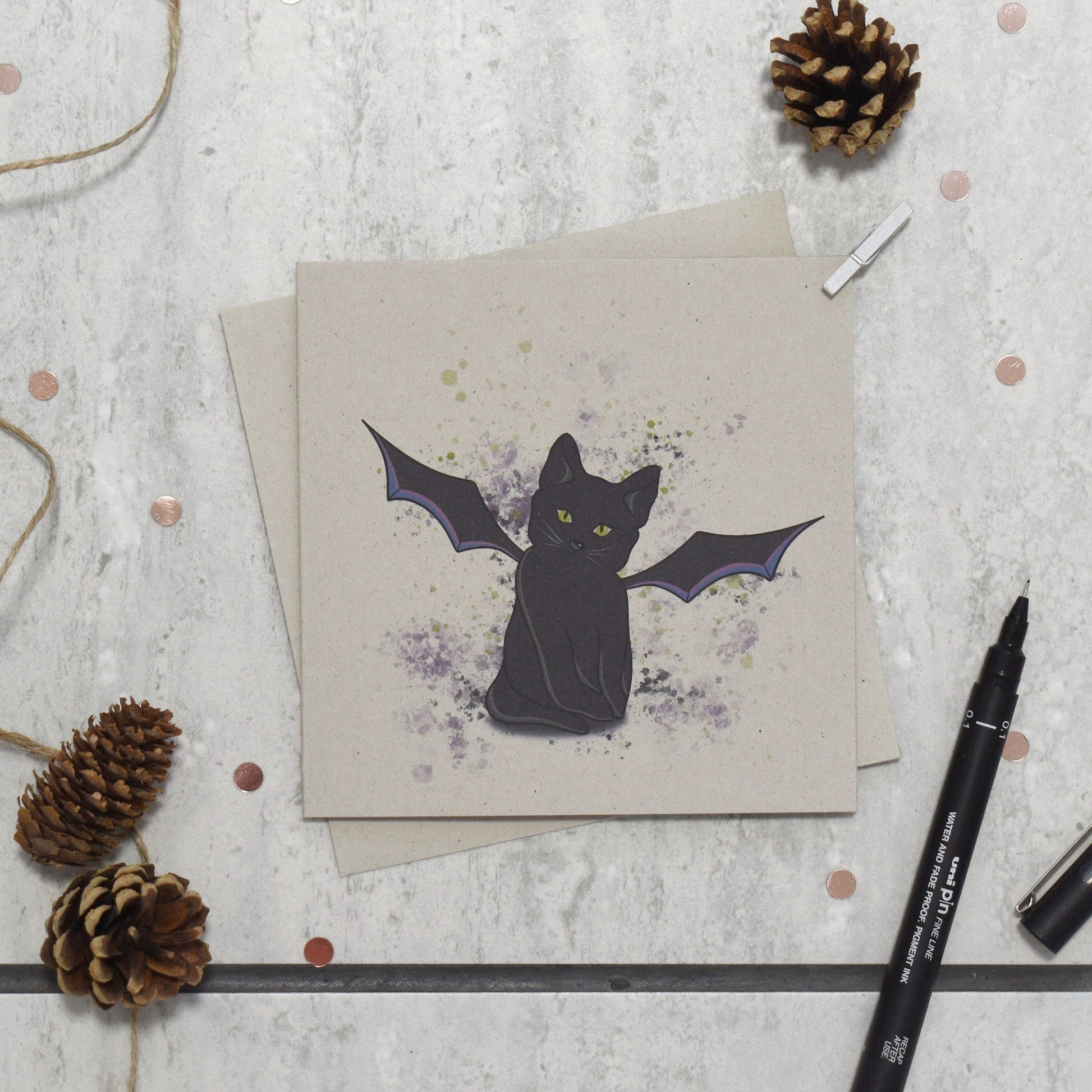 Greeting card with a black cat illustration on a textured surface with pine cones and markers.