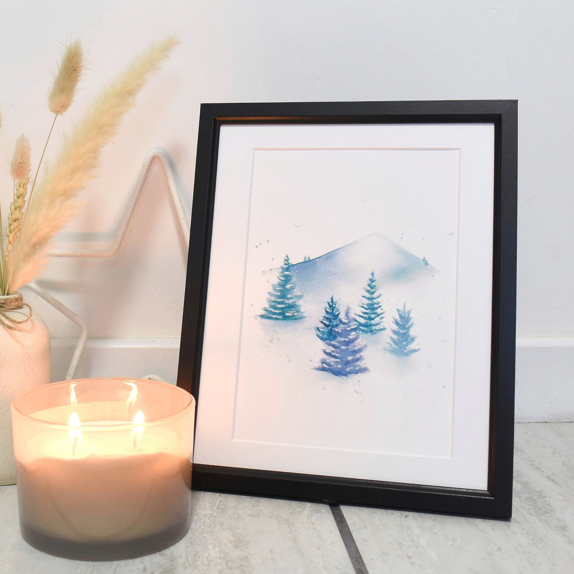 Framed artwork of trees and mountains next to a lit candle and vase with dried plants on a light wooden floor.