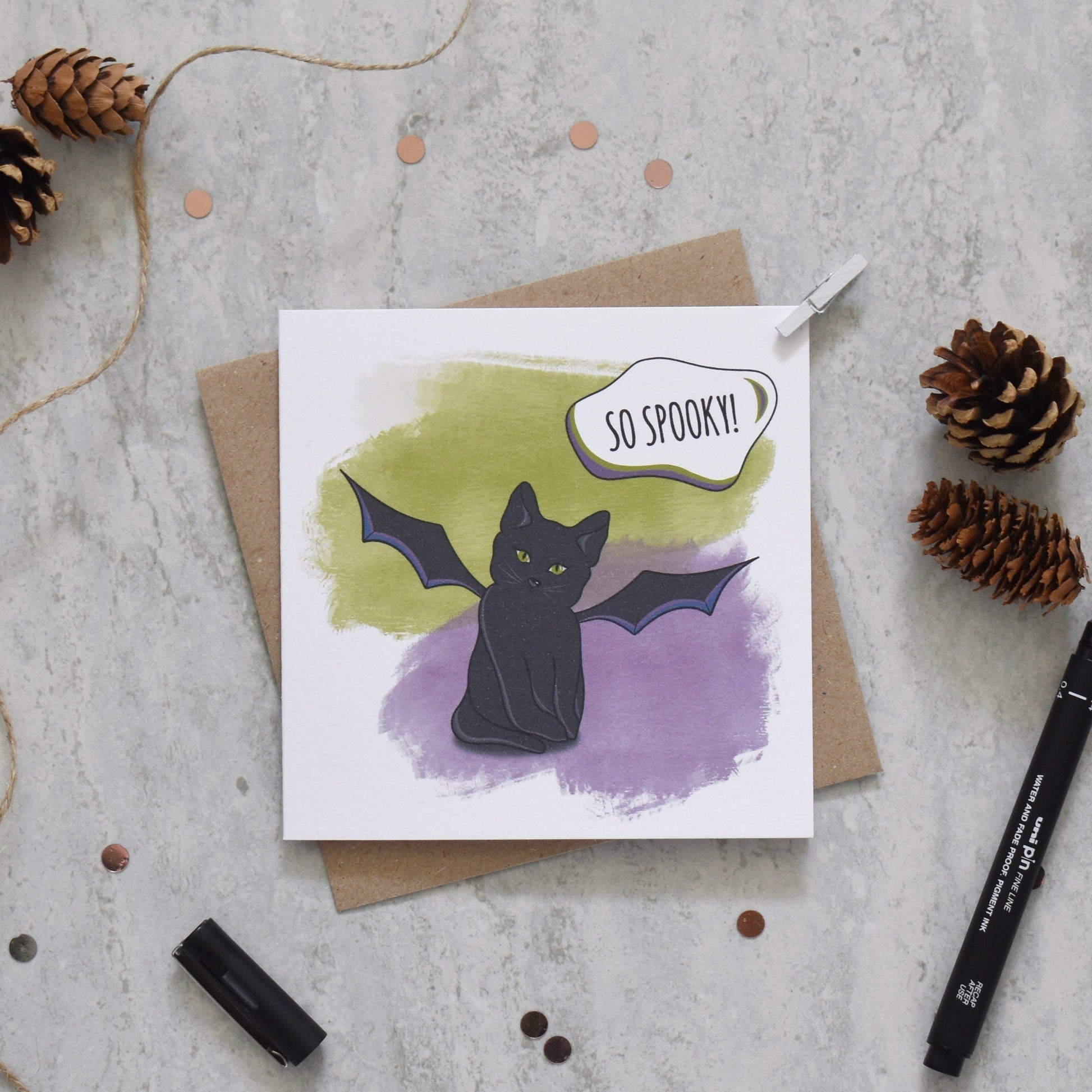 Greeting card with a black cat illustration and 'So Spooky!' text, surrounded by decorative items on a textured surface.