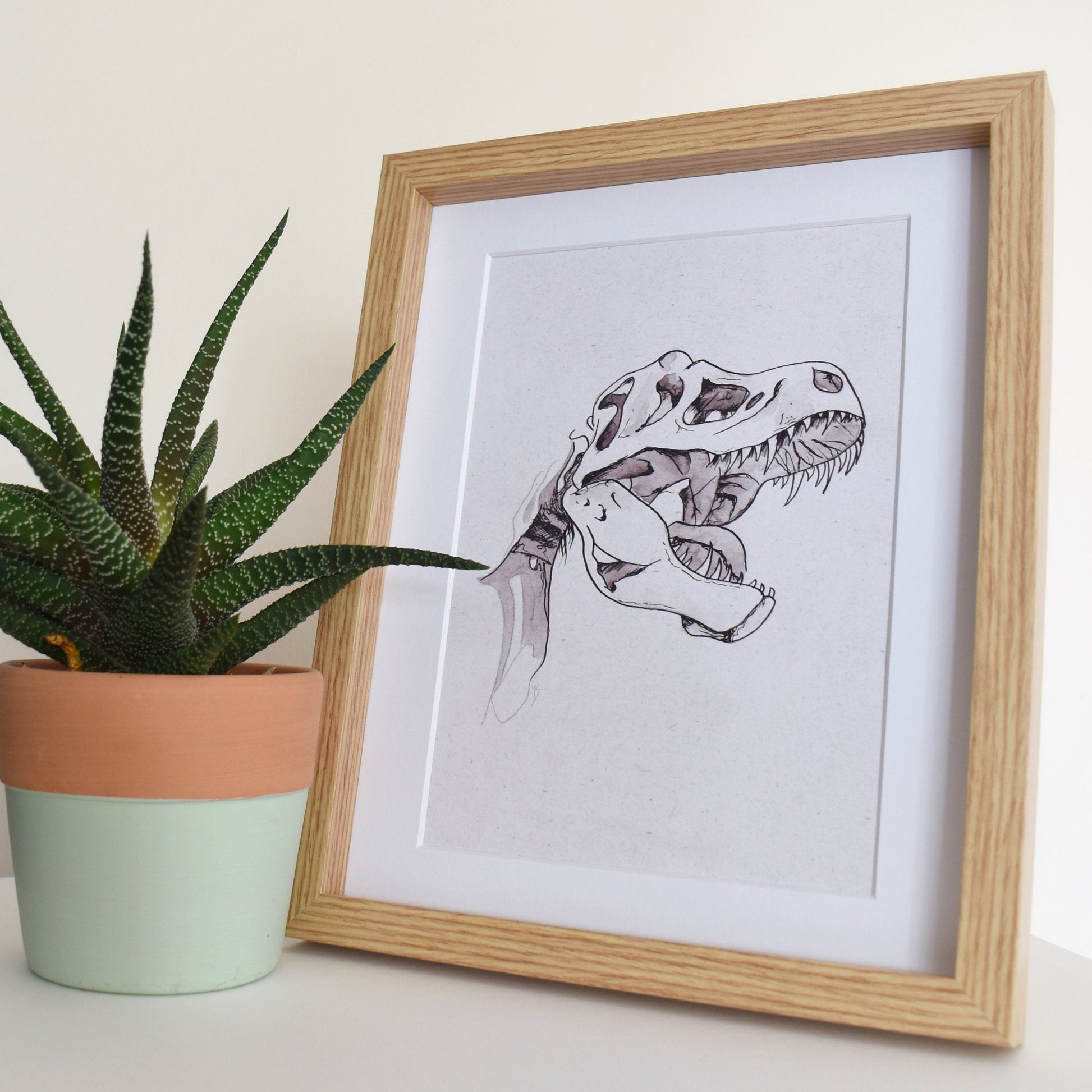 Framed artwork of a dinosaur next to a potted plant on a white surface.