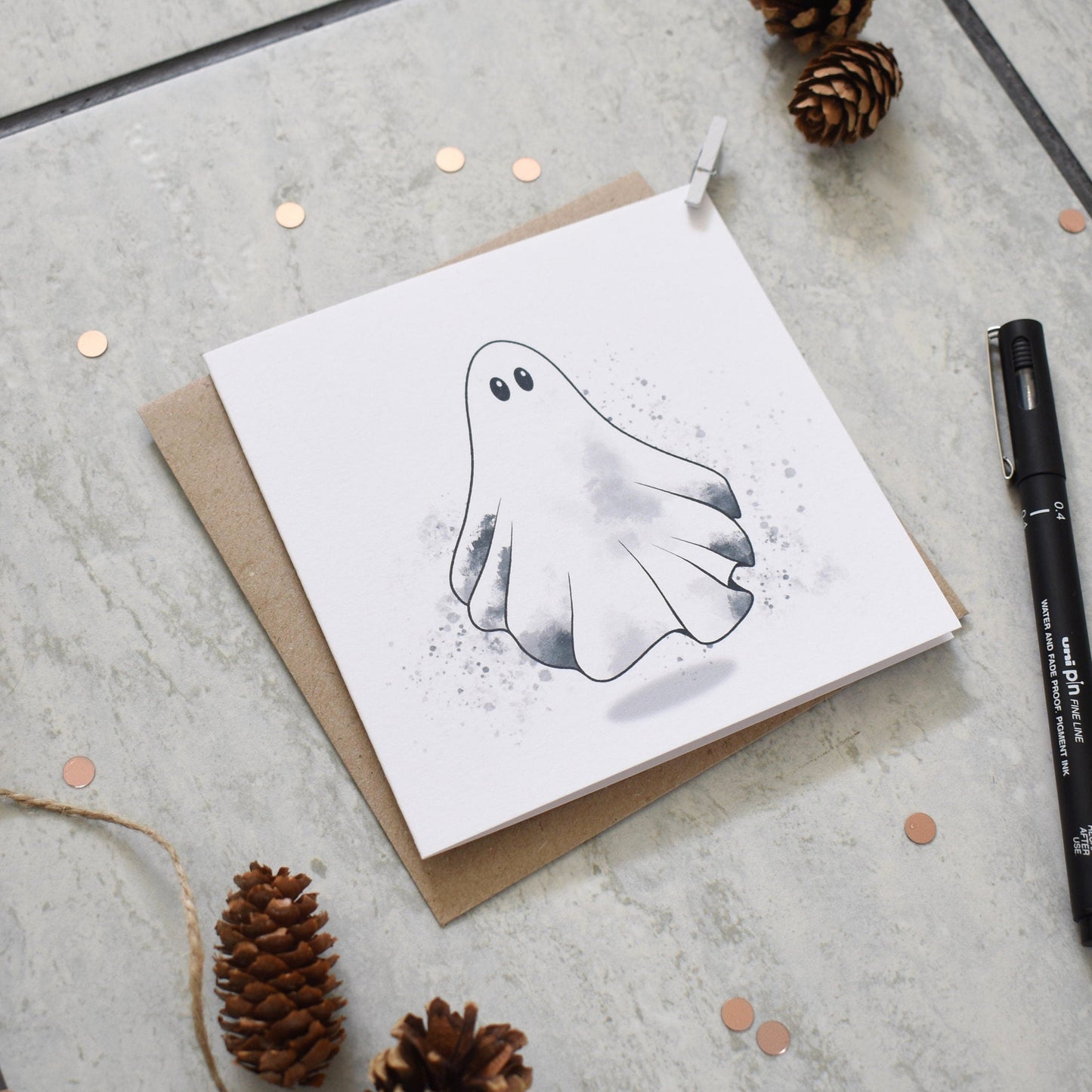 Greeting card with ghost illustration on a textured surface with pine cones and a pen.