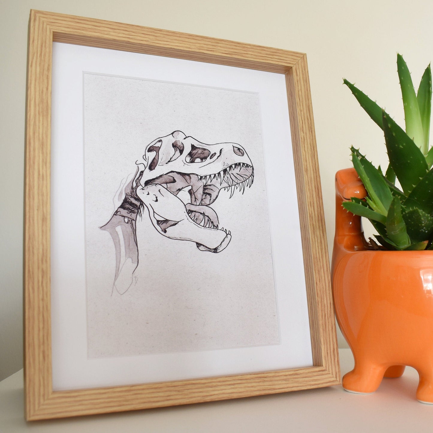 Framed dinosaur illustration next to an orange planter with a plant on a neutral background