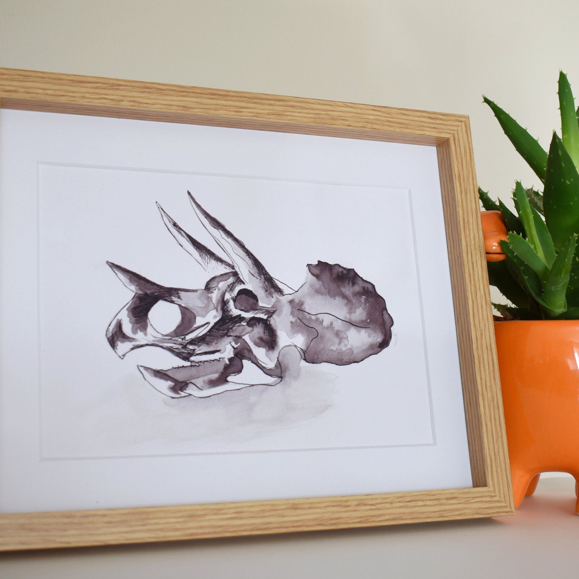 Framed artwork of a triceratops skull next to an orange vase with a plant on a neutral background