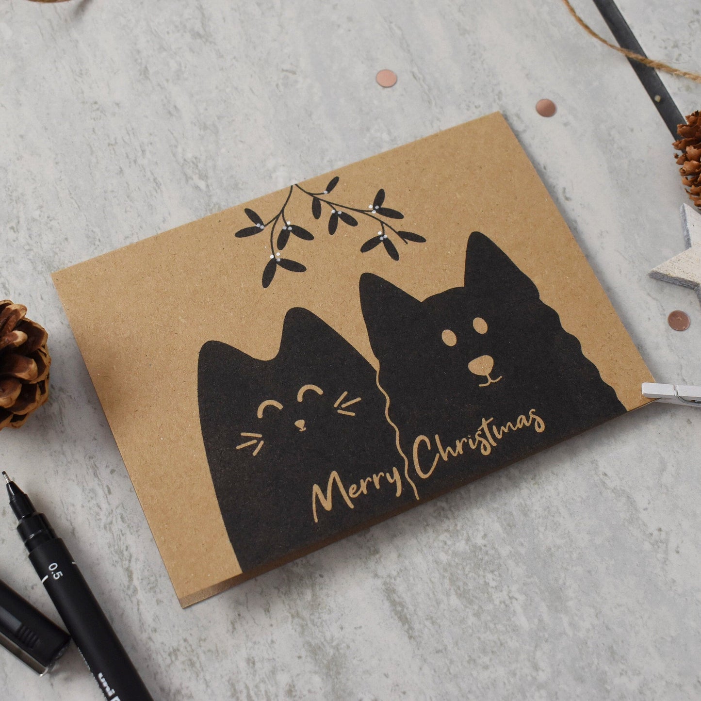 Kraft paper card with black cat illustration and 'Merry Christmas' text on a gray surface with decorative elements.