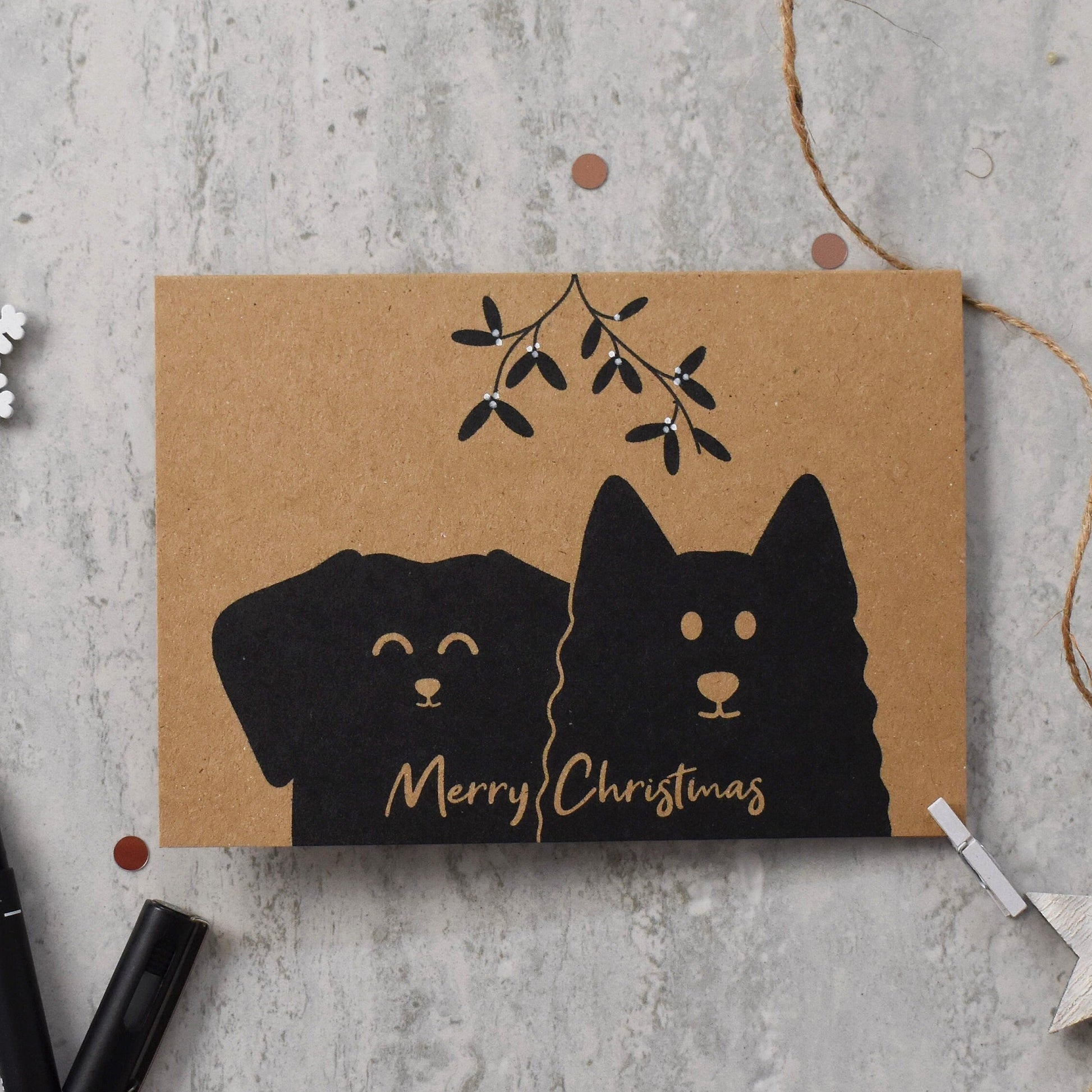 Kraft paper card with black dog illustration and 'Merry Christmas' text on a gray surface.