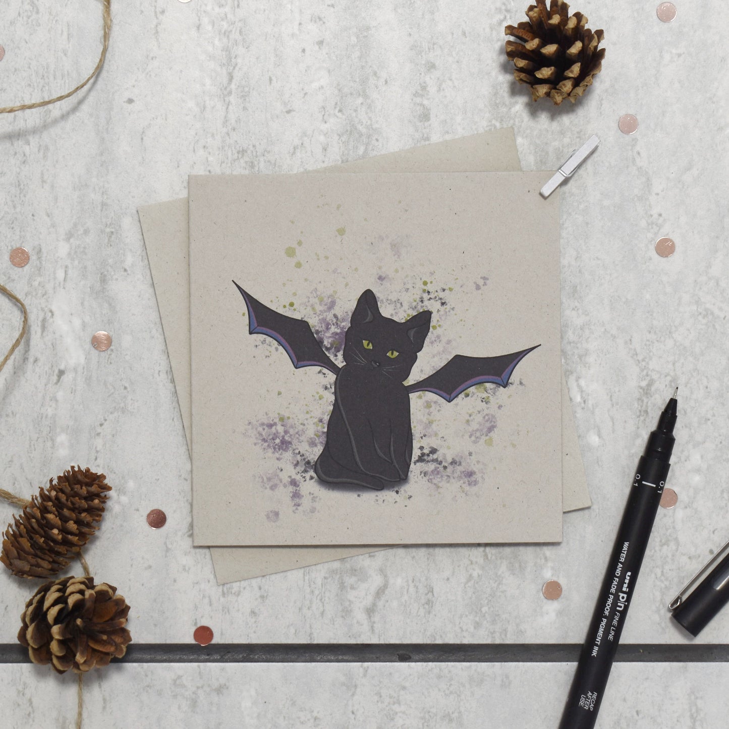 Greeting card with a black cat illustration on a textured surface with pine cones and a pen.