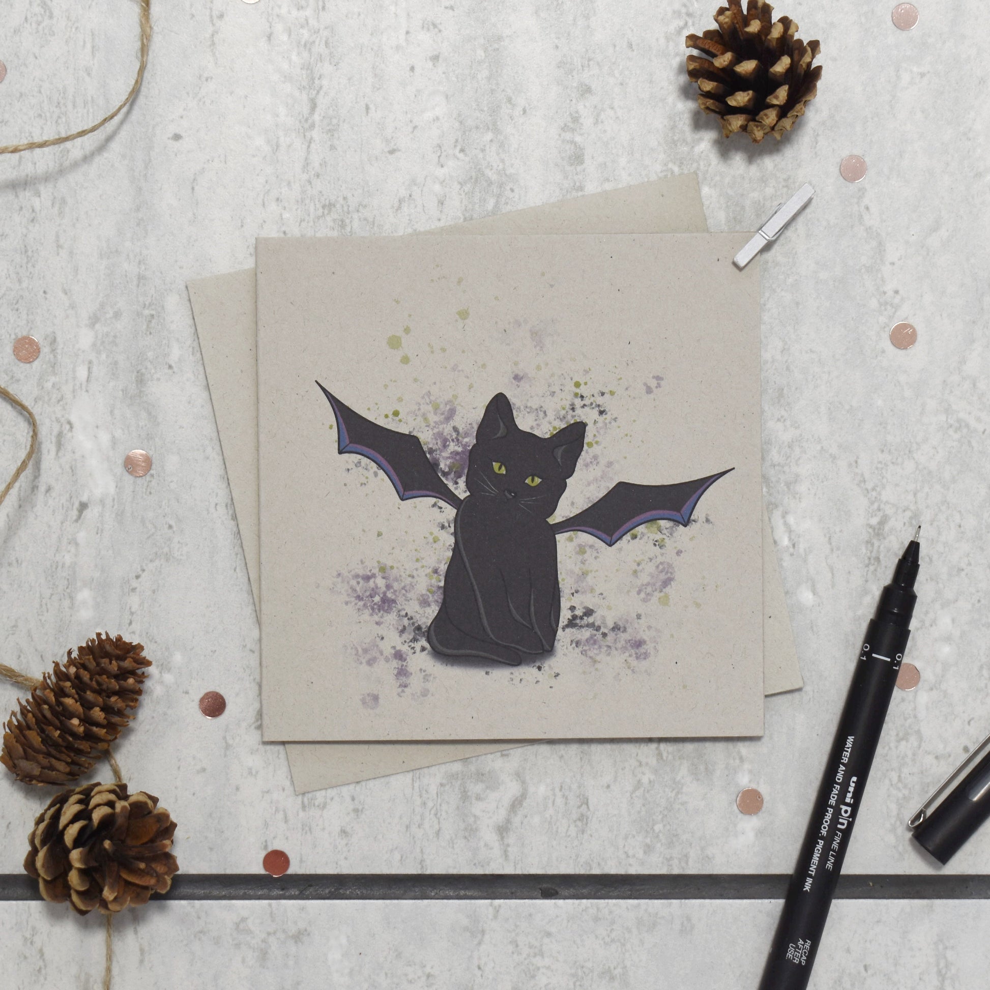 Greeting card with a black cat illustration on a textured surface with pine cones and a pen.