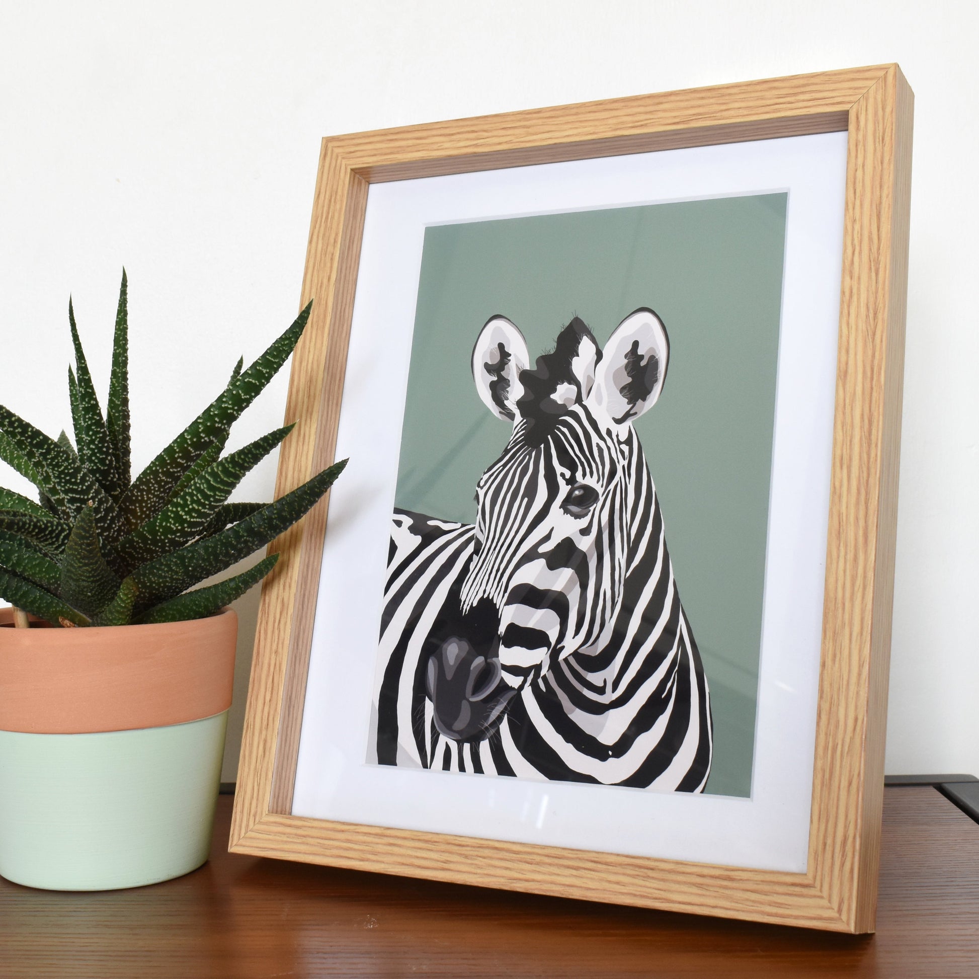 Framed picture of a zebra next to a potted plant on a wooden surface.
