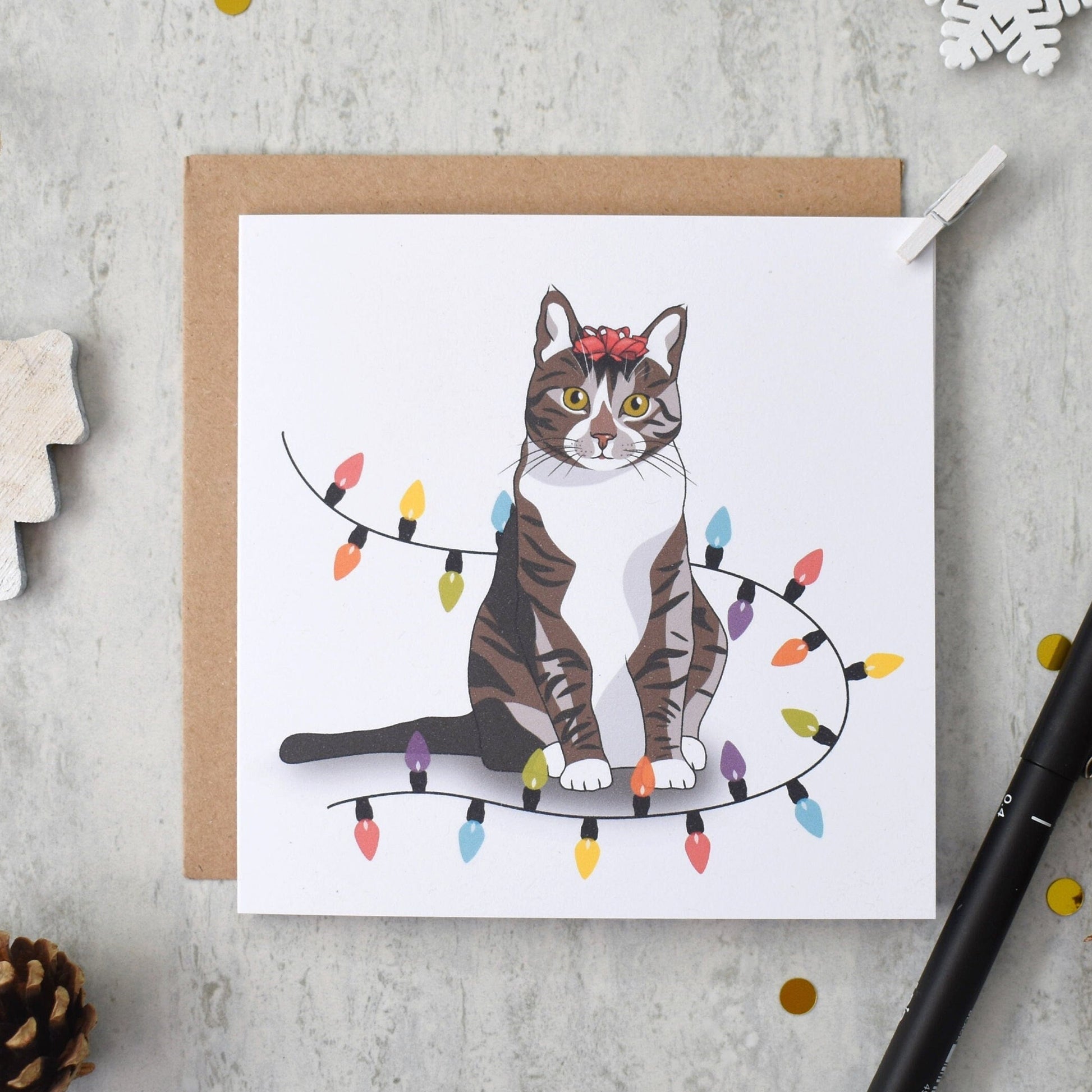 Card with a cat illustration on a string of lights, surrounded by decorative items on a gray surface.