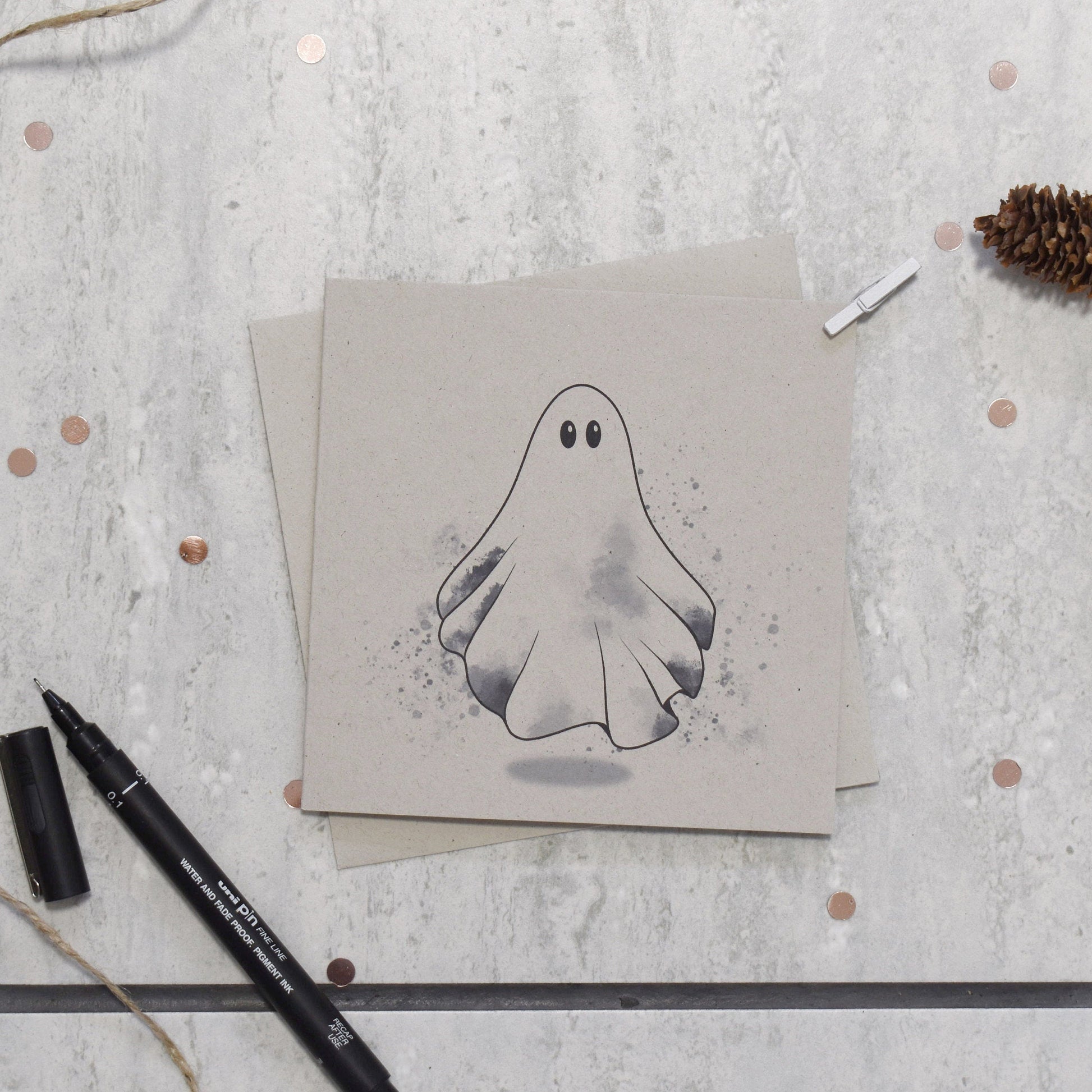 Card with a ghost illustration on a textured surface with pine cones and a pen.